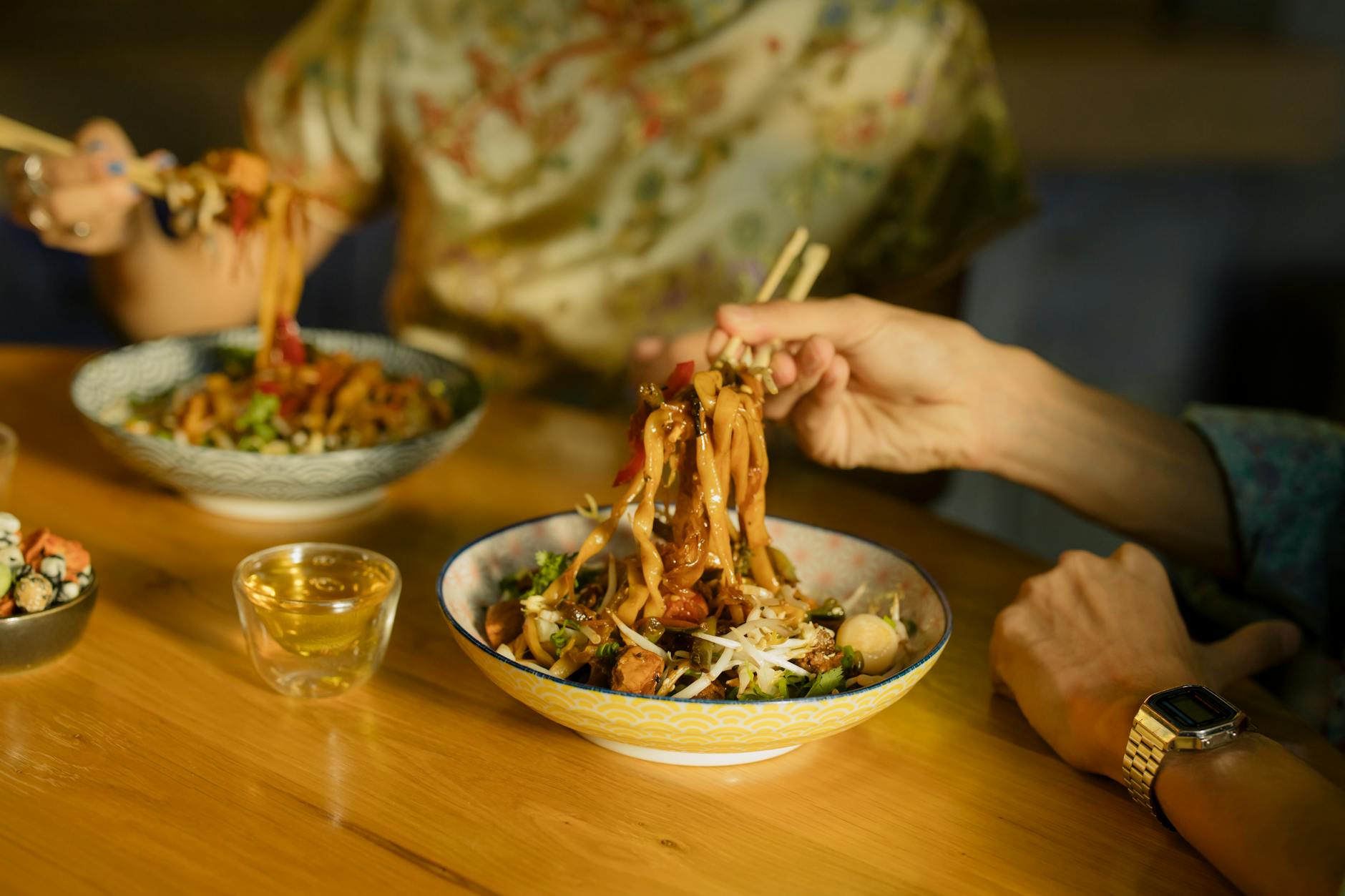 Two diners with chopsticks lifting noodles from shared bowls on a wooden table