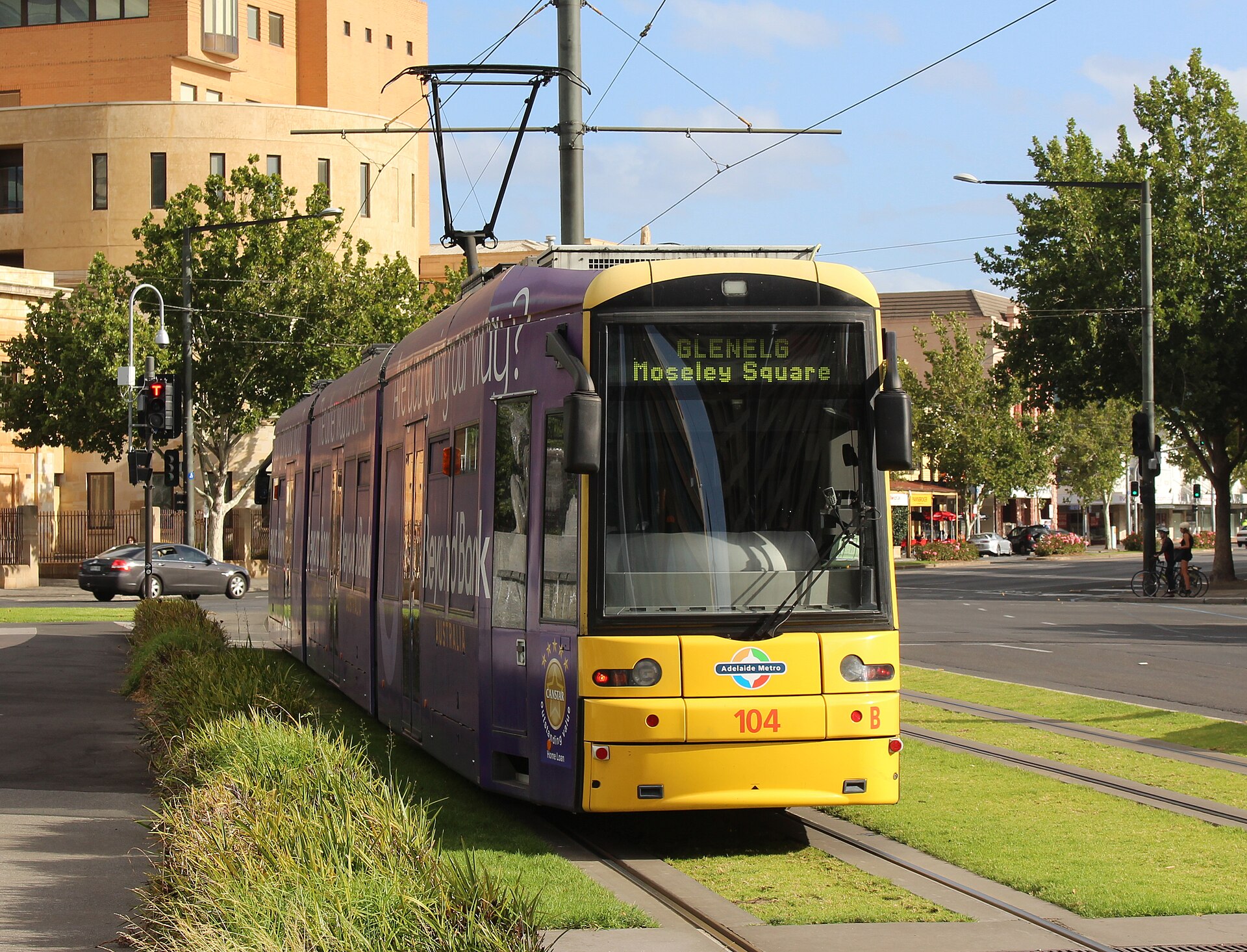 An Adelaide Metro tram bound for Moseley Square in Glenelg travelling along a grassed track beside a road