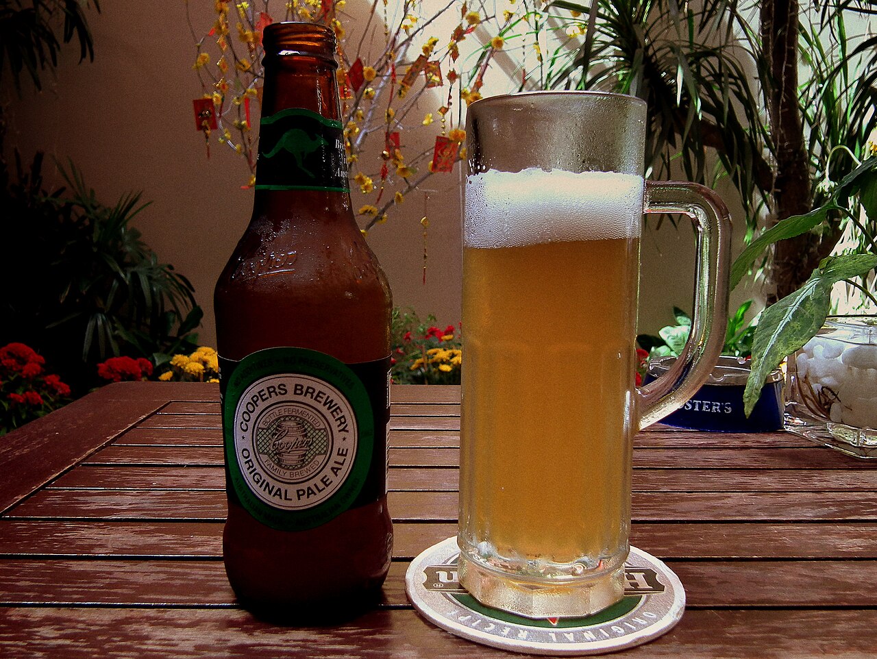 A brown bottle of Coopers Original Pale Ale beside a tall glass of cloudy golden beer on a wooden table