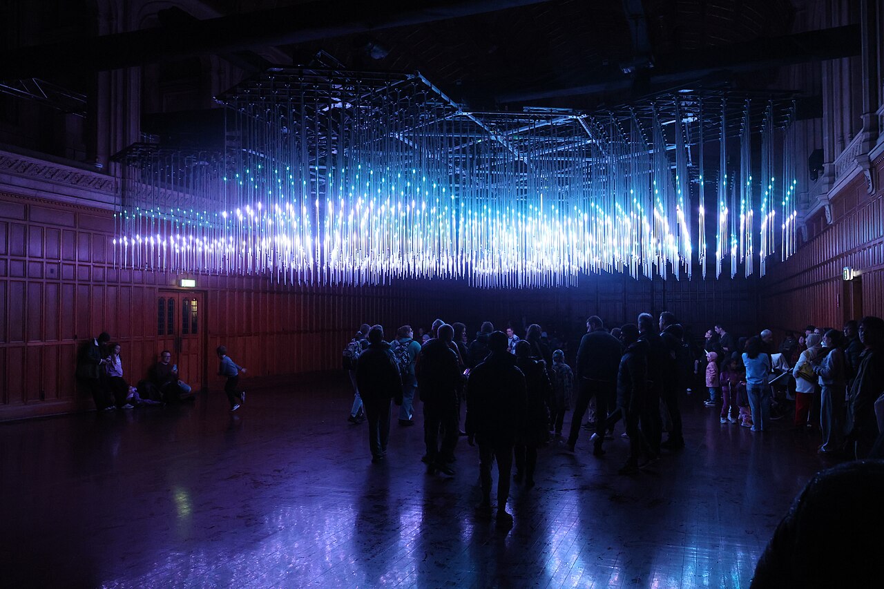 Visitors standing beneath a ceiling of hanging vertical light rods glowing blue and purple inside Bonython Hall