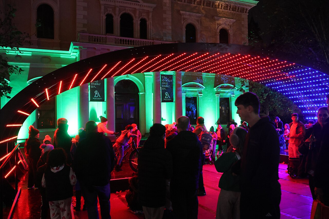 Crowds passing under a curved arch of red and blue LED lights in front of the State Library of South Australia, its facade lit green