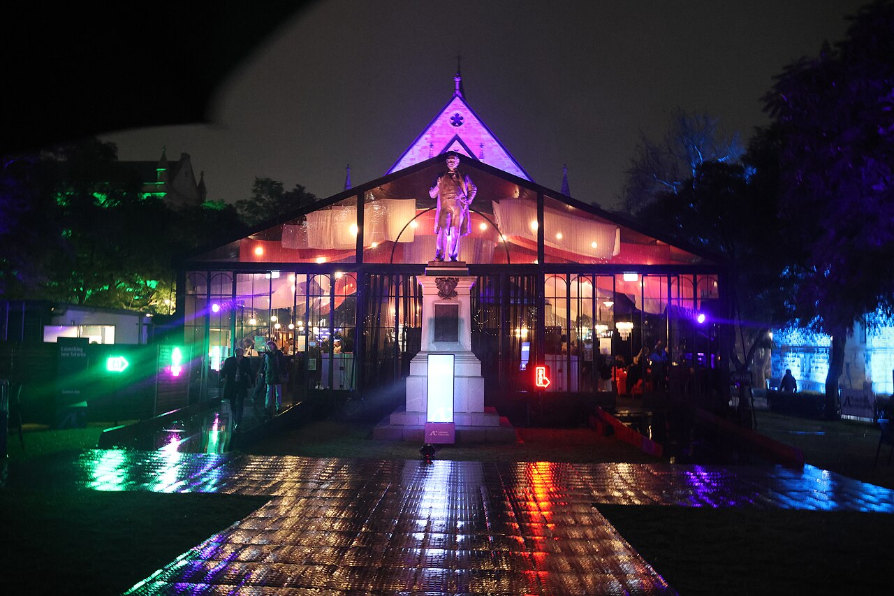 A glass pavilion on the University of Adelaide campus glowing pink and purple at night, with coloured light reflecting on wet paving