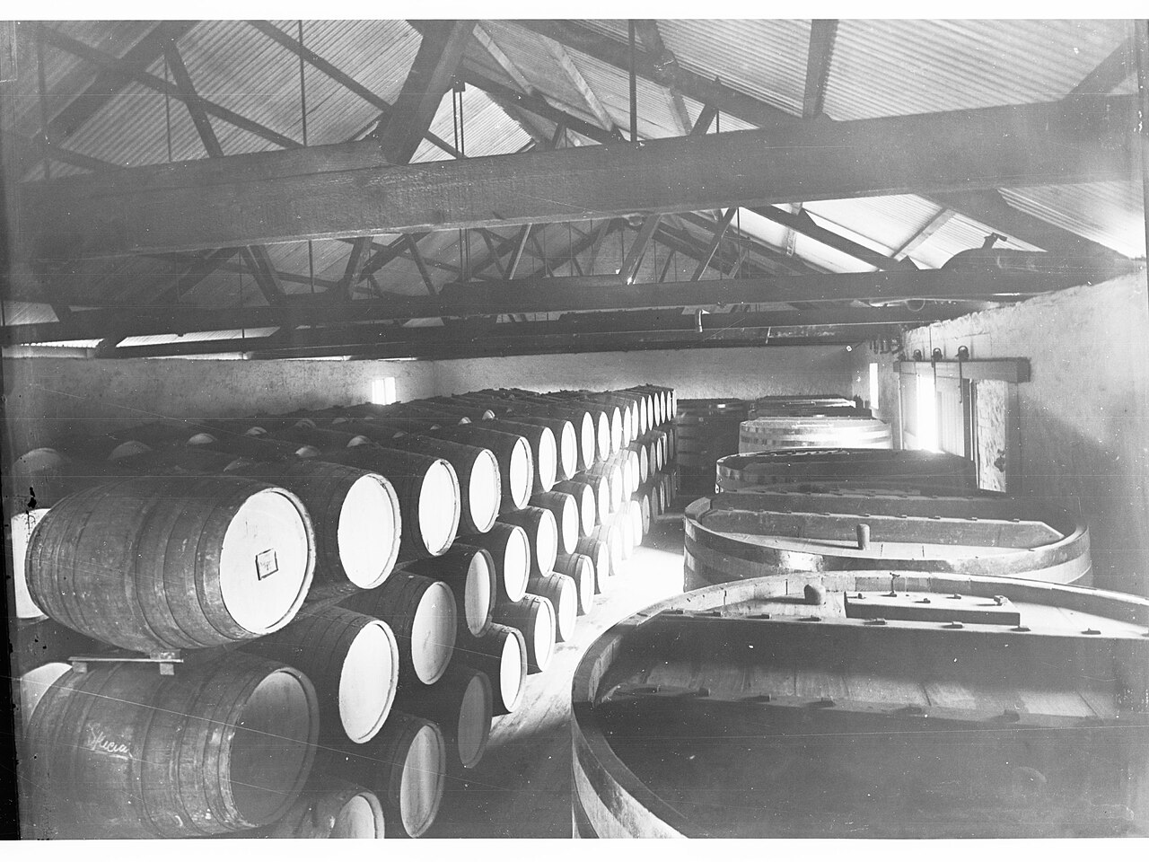 A black-and-white photograph of barrels and large open vats stacked inside the Penfold wine cellars at Magill around 1915