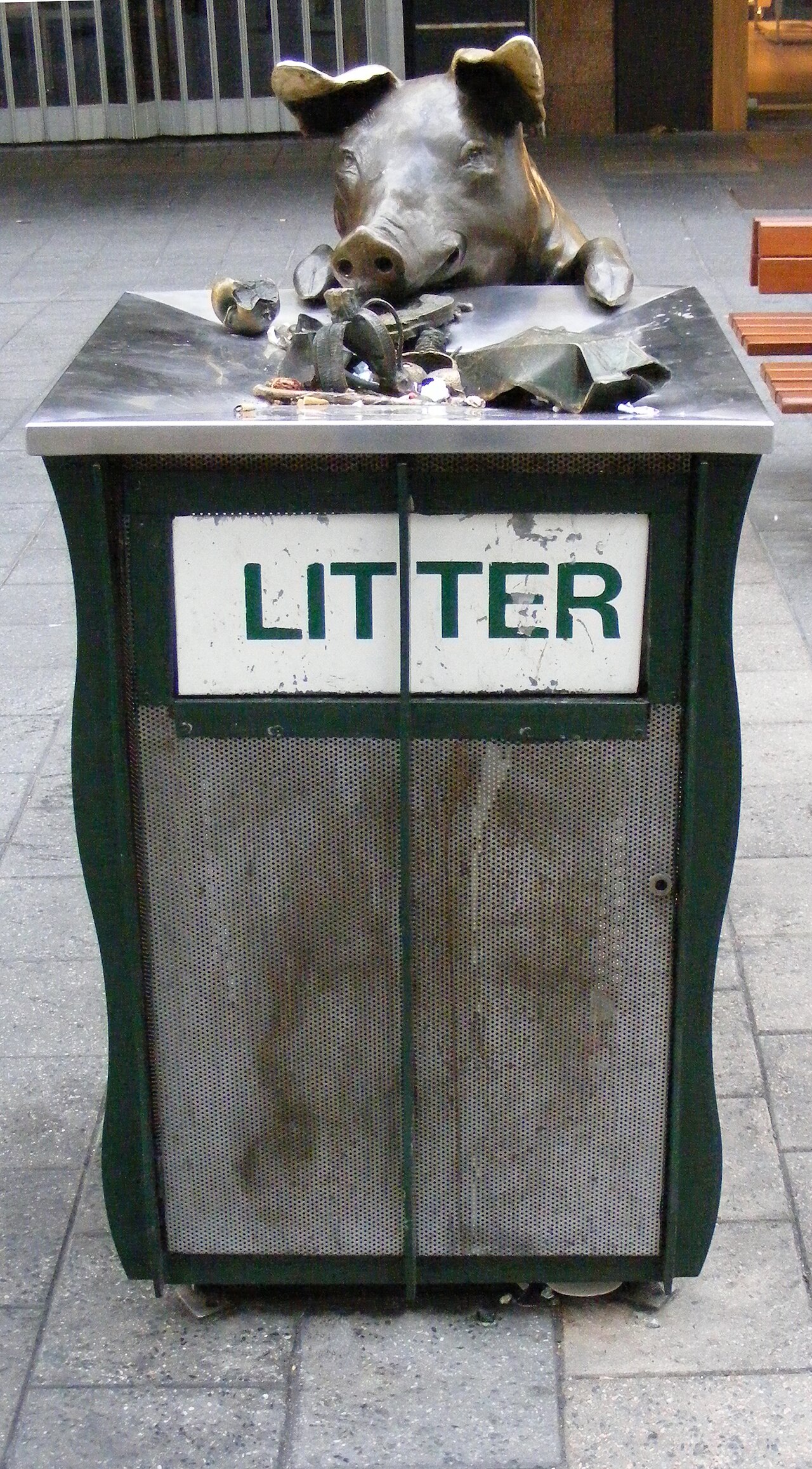 A bronze pig with its snout and front trotters over the lid of a green litter bin, surrounded by bronze scraps of rubbish