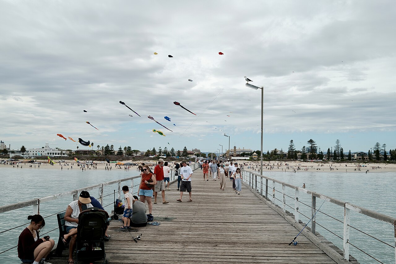 Dozens of colourful kites flying above Semaphore jetty with people walking along the boardwalk and the beach behind