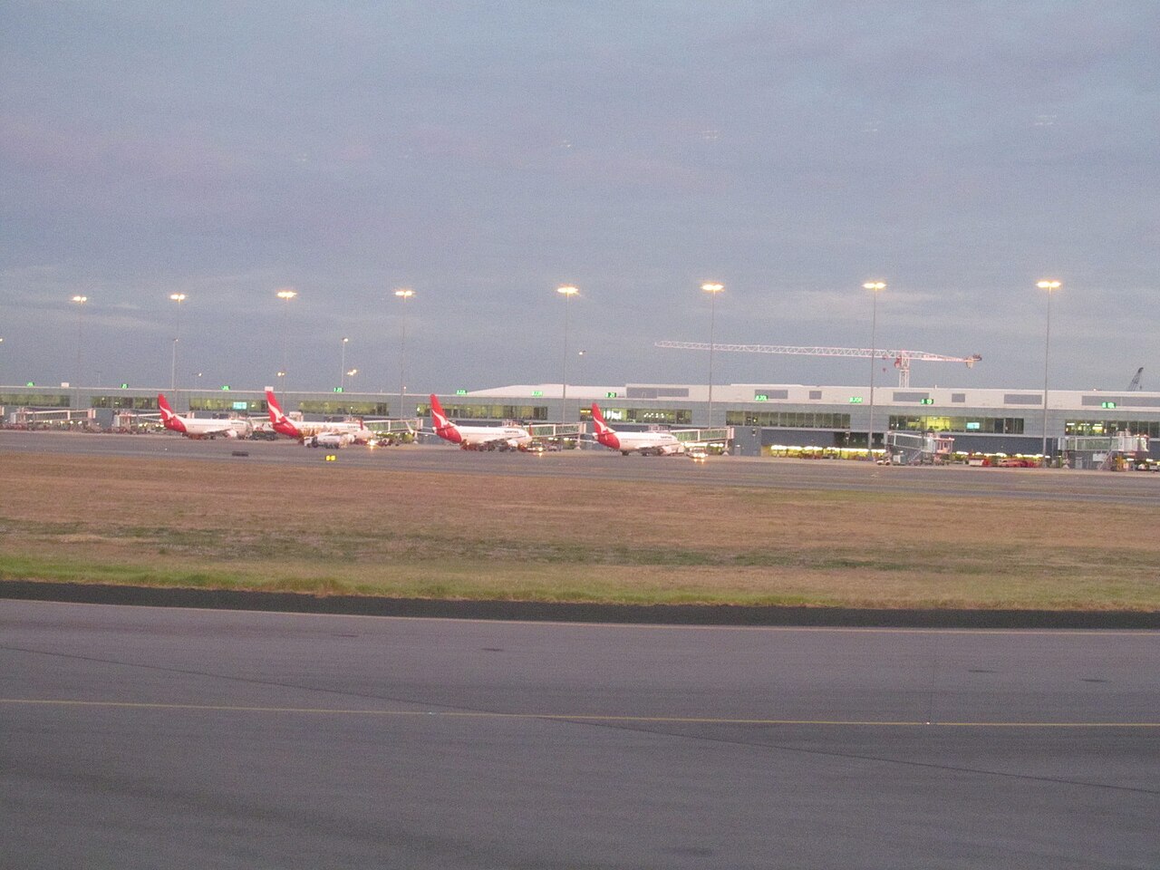 Four Qantas jets parked at the aerobridges of Adelaide Airport's long, low terminal at dawn, seen across a grassed area and taxiway, with a construction crane behind