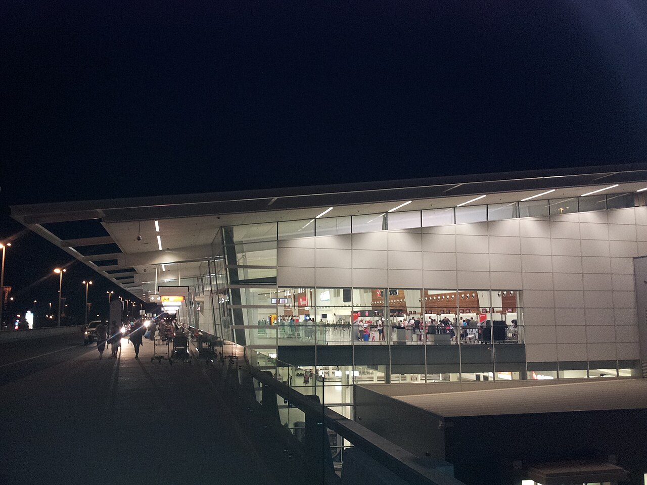 The front of Adelaide Airport's terminal at night, with the elevated departures road and footpath on the left and check-in counters lit up behind tall glass windows