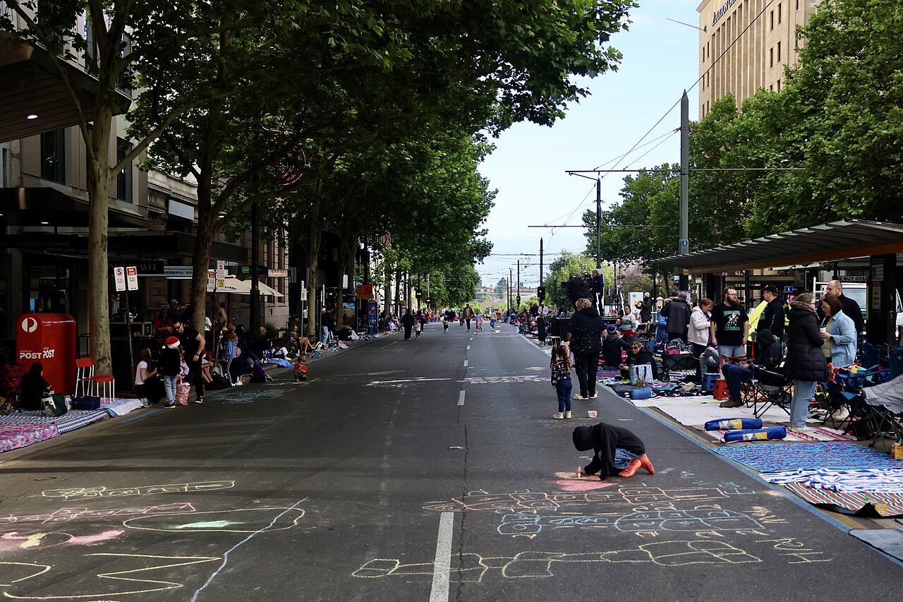 King William Street in Adelaide early on Pageant morning, with families camped along both kerbs on rugs and chairs and chalk drawings covering the road