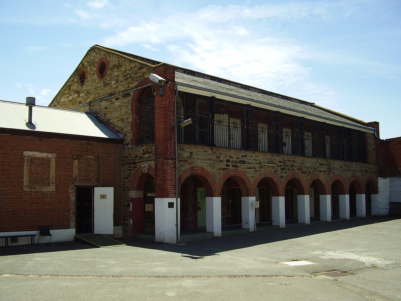 A two-storey cell block of stone and red brick at the Adelaide Gaol, with an arched arcade along the ground floor and a paved yard in front