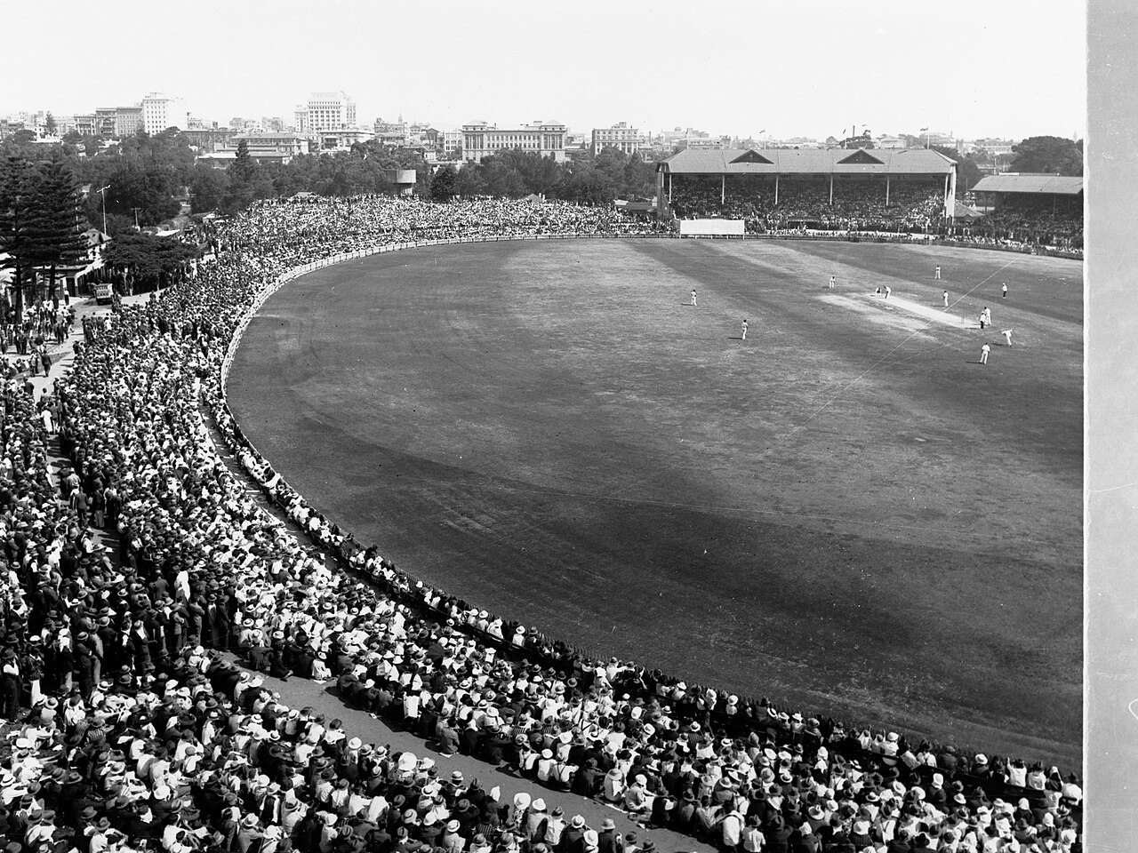 Black-and-white photograph of a huge crowd encircling the field at Adelaide Oval during the Fourth Test between Australia and England in 1937, with the city beyond