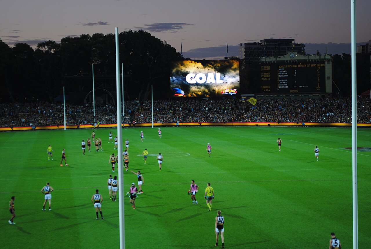 A night AFL match at Adelaide Oval with players spread across the floodlit ground and the word GOAL on the big screen behind the goalposts