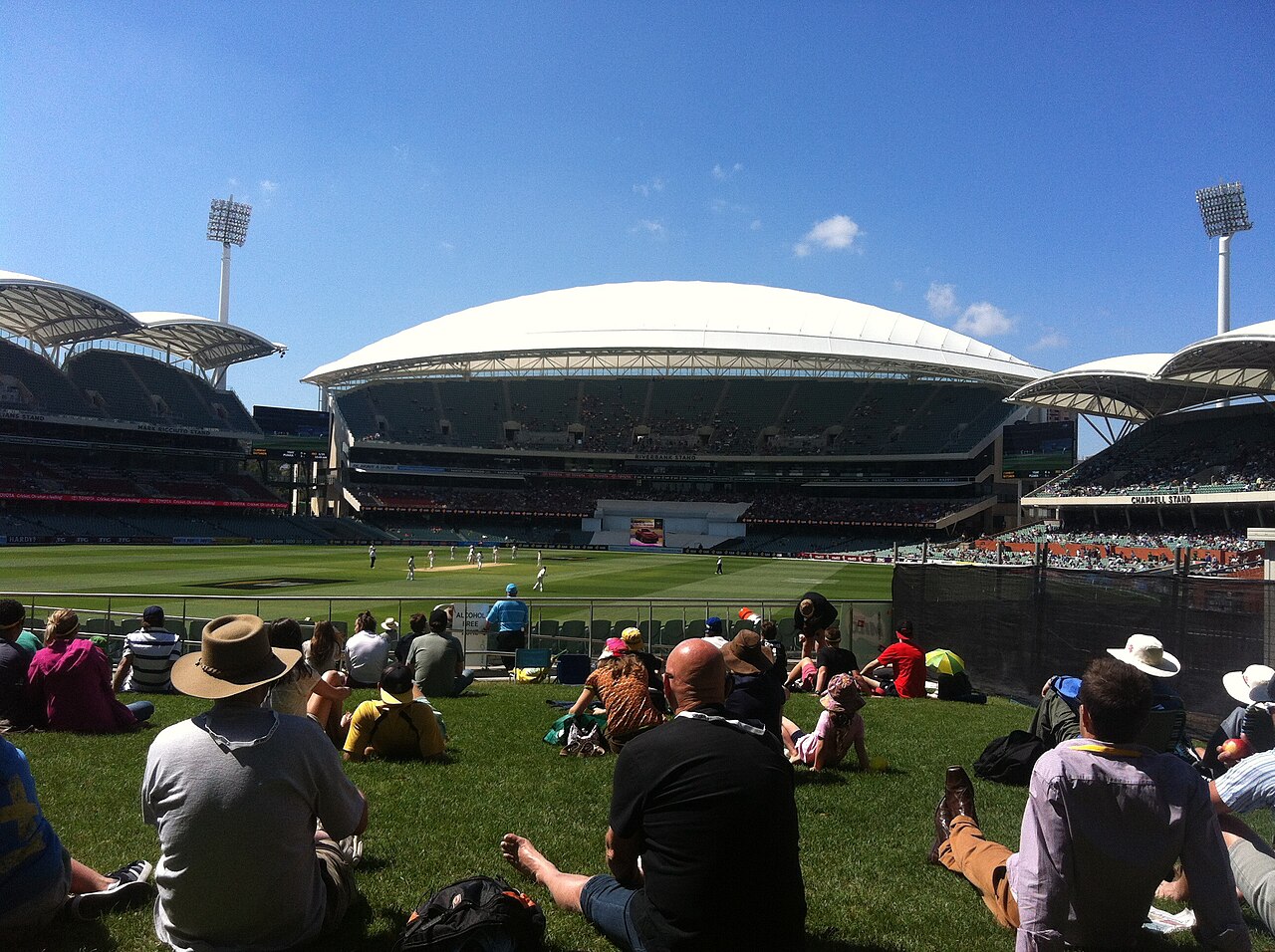 Spectators sitting on the grass of the Hill at Adelaide Oval during a Test match, looking across the ground to the modern grandstands with their white roof canopies