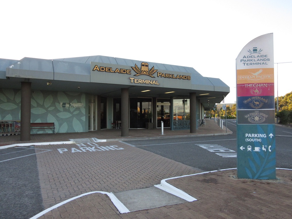The entrance of Adelaide Parklands Terminal, a low building with a curved grey roof, timber bench and paved forecourt, beside a tall sign listing the long-distance trains that use it