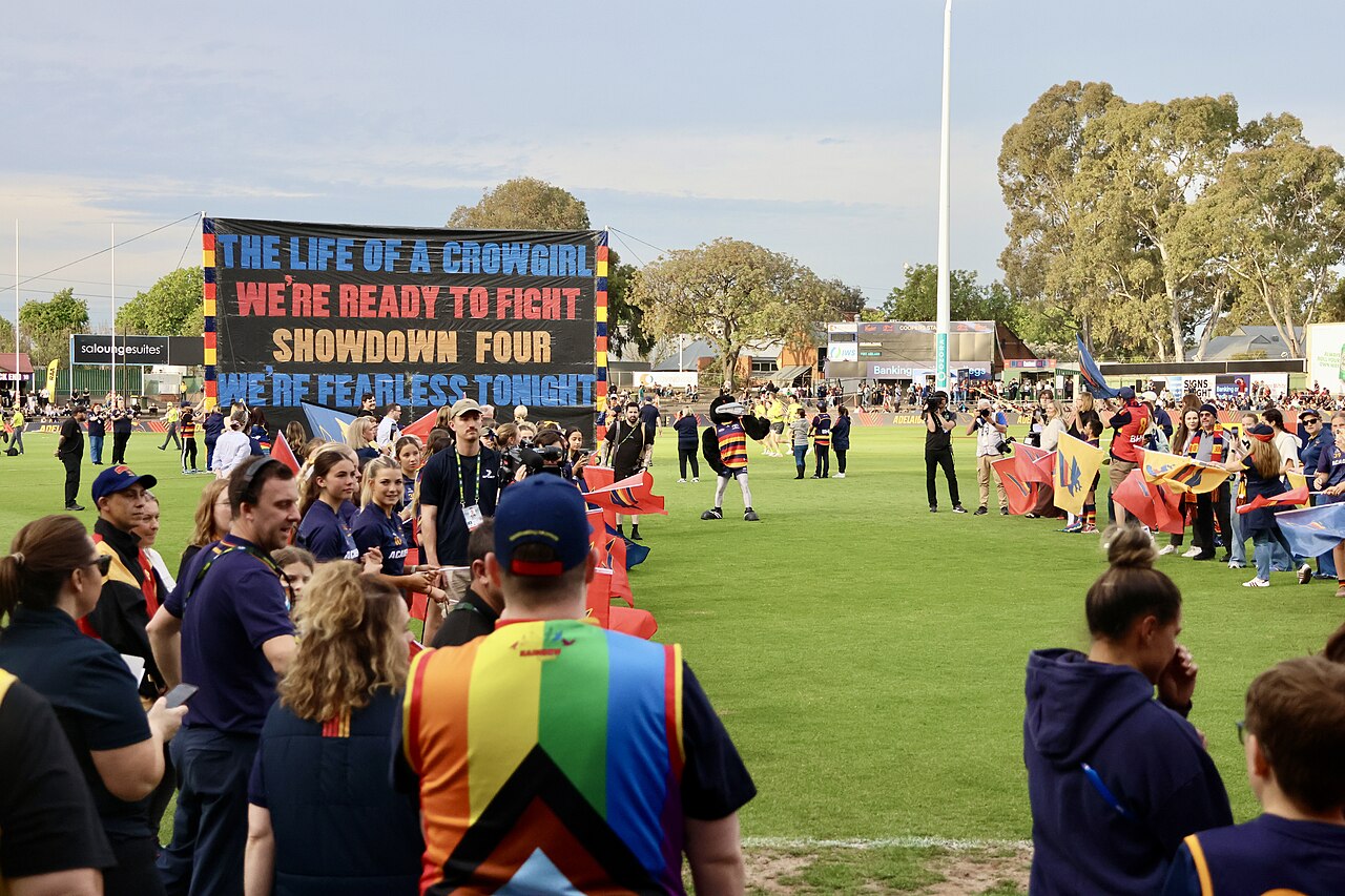 Crows supporters forming a tunnel with flags at Norwood Oval as the Adelaide AFLW team runs out for a Showdown, under a banner reading Showdown Four
