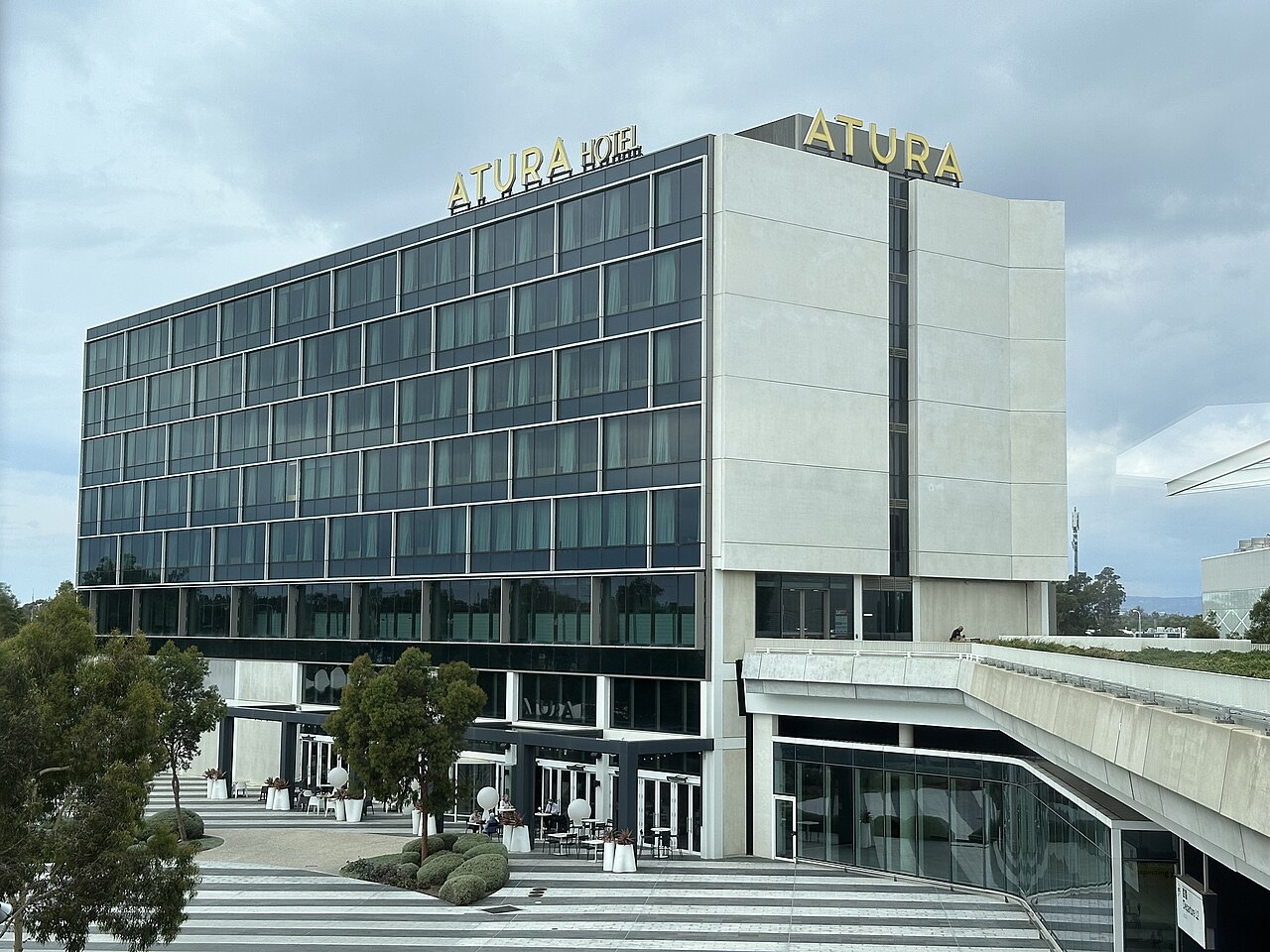 The Atura Adelaide Airport hotel, a modern multi-storey building with a grid of dark windows and a pale stone tower, beside the terminal walkway