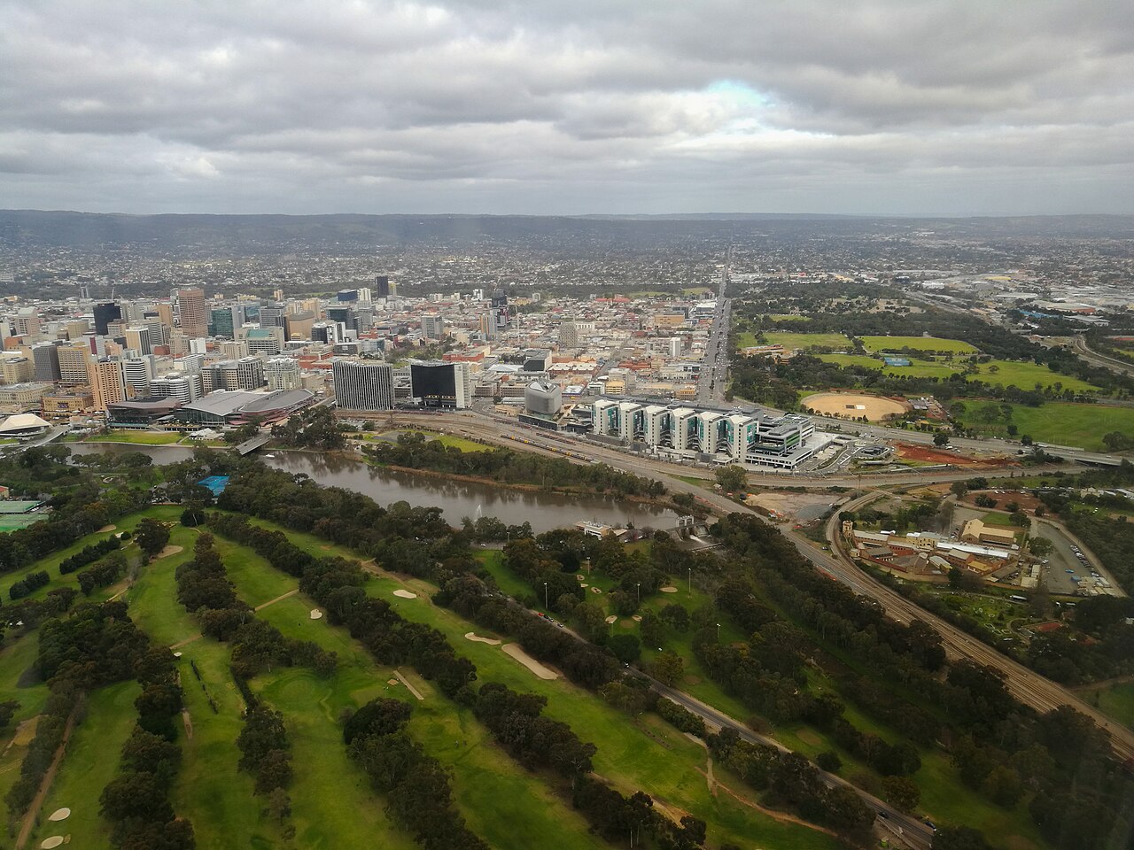 Aerial view looking east over Bonython Park and the River Torrens parklands towards the Adelaide city skyline, with the Adelaide Hills on the horizon