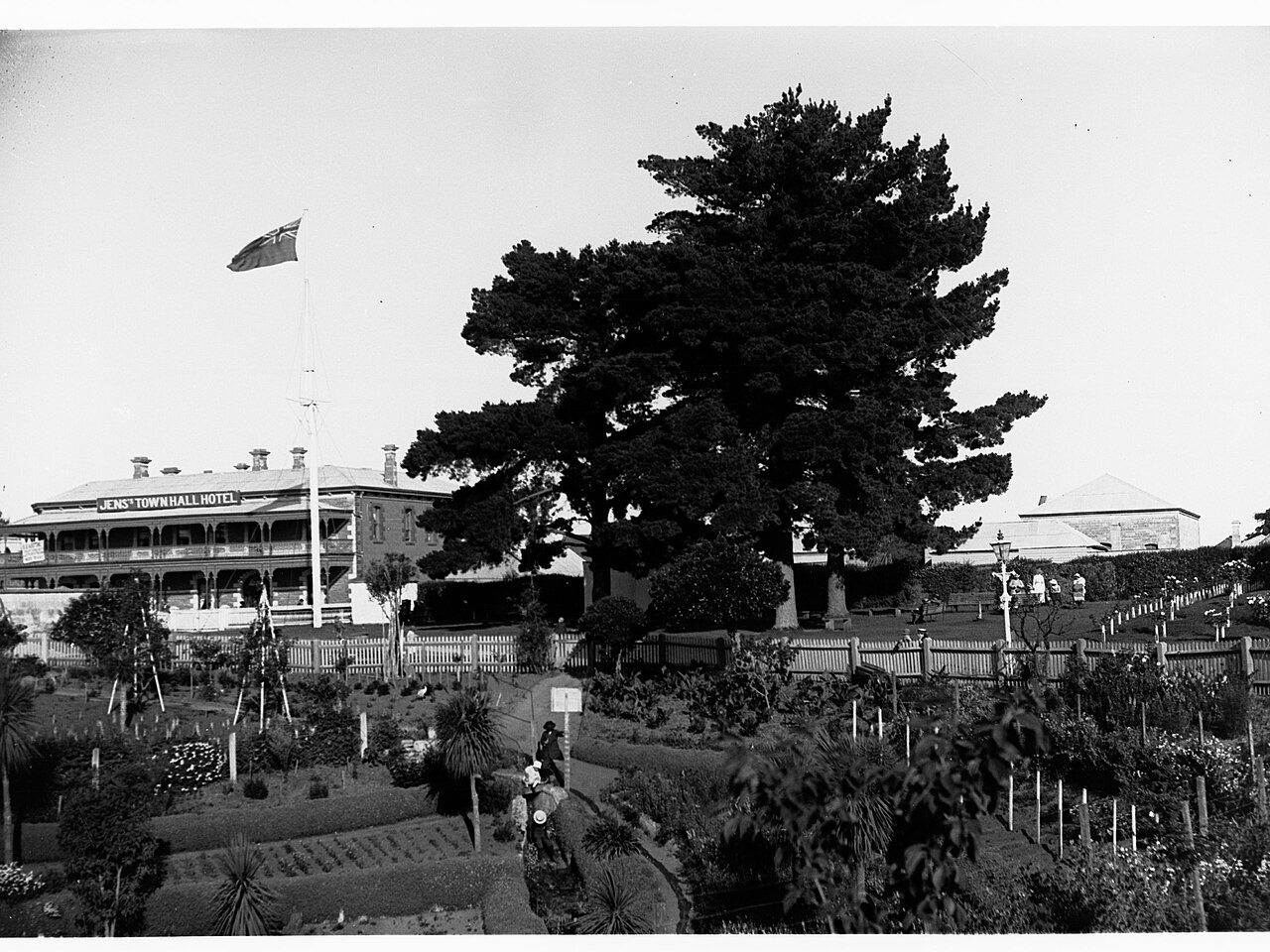 A black-and-white archive photograph of the Cave Garden in Mount Gambier, with flower beds and paths in front of a large tree and a two-storey hotel building with a flag flying above it