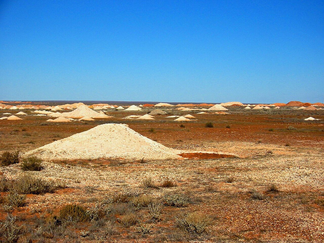 Pale spoil heaps from opal mining shafts scattered across flat red-brown scrubland near Coober Pedy under a deep blue sky