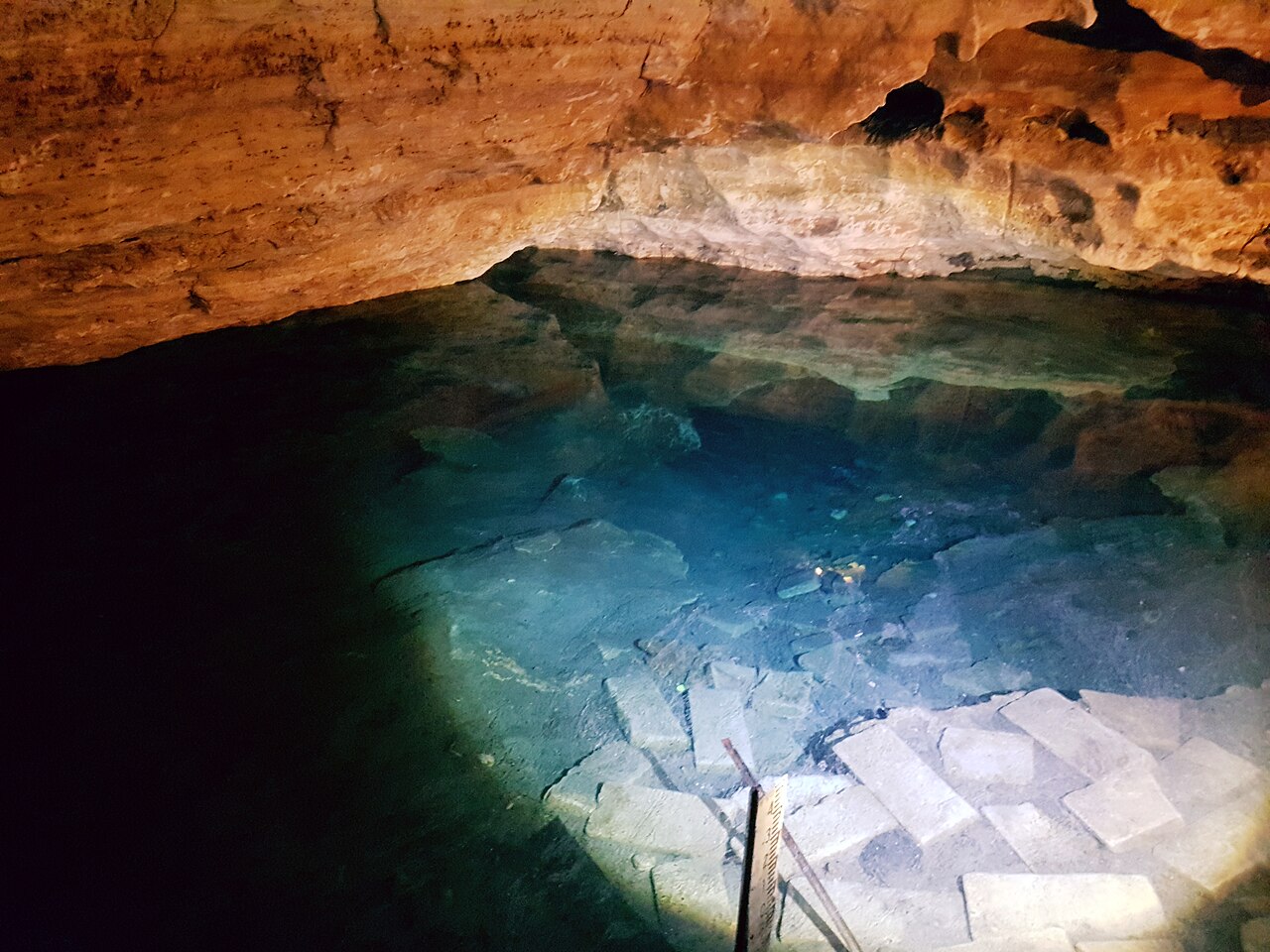 Clear blue-green water filling the floor of Engelbrecht Cave beneath a low, rust-coloured limestone ceiling, with submerged stone steps visible below the surface