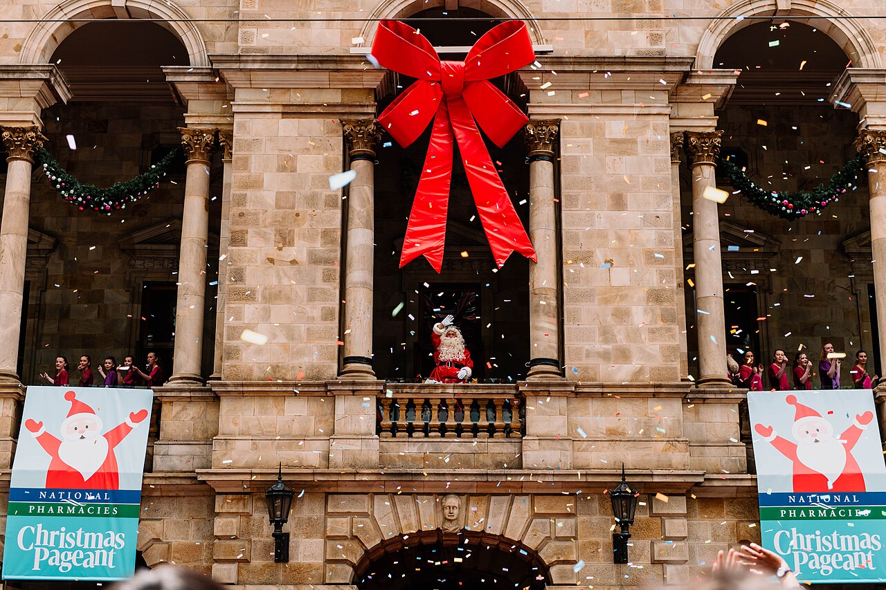 Father Christmas waving from the balcony of the Adelaide Town Hall beneath a giant red bow, with confetti falling and Christmas Pageant banners on either side