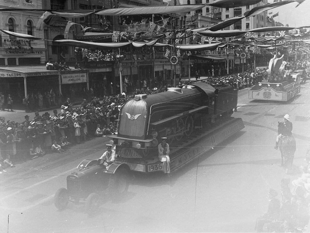 Black-and-white photograph of a streamlined locomotive float driving up King William Street in the 1936 John Martin's Christmas Pageant, with large crowds and decorations strung overhead