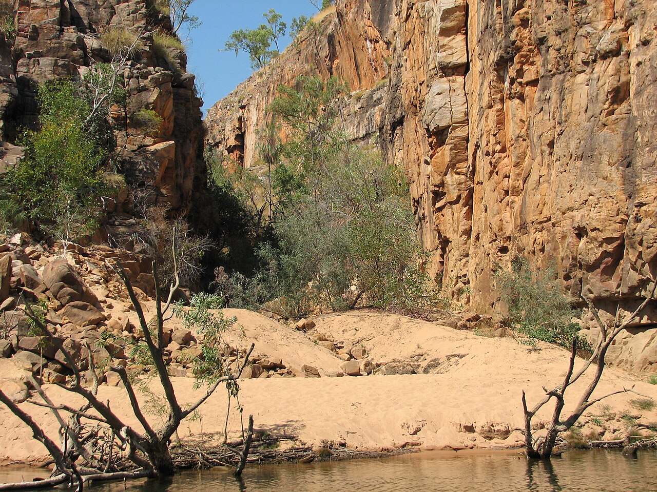 Tall orange sandstone cliffs rising above a sandy bank and calm water in Katherine Gorge, with scrubby trees growing among the rocks