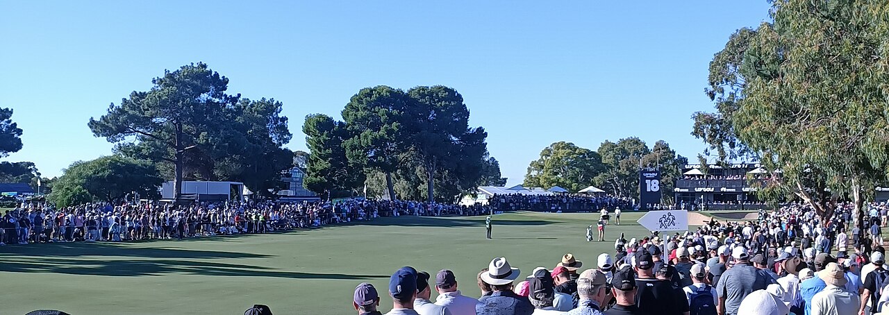 A large crowd of golf fans packed around the 18th green at The Grange Golf Club during LIV Golf Adelaide 2023, with tall pines and grandstands behind