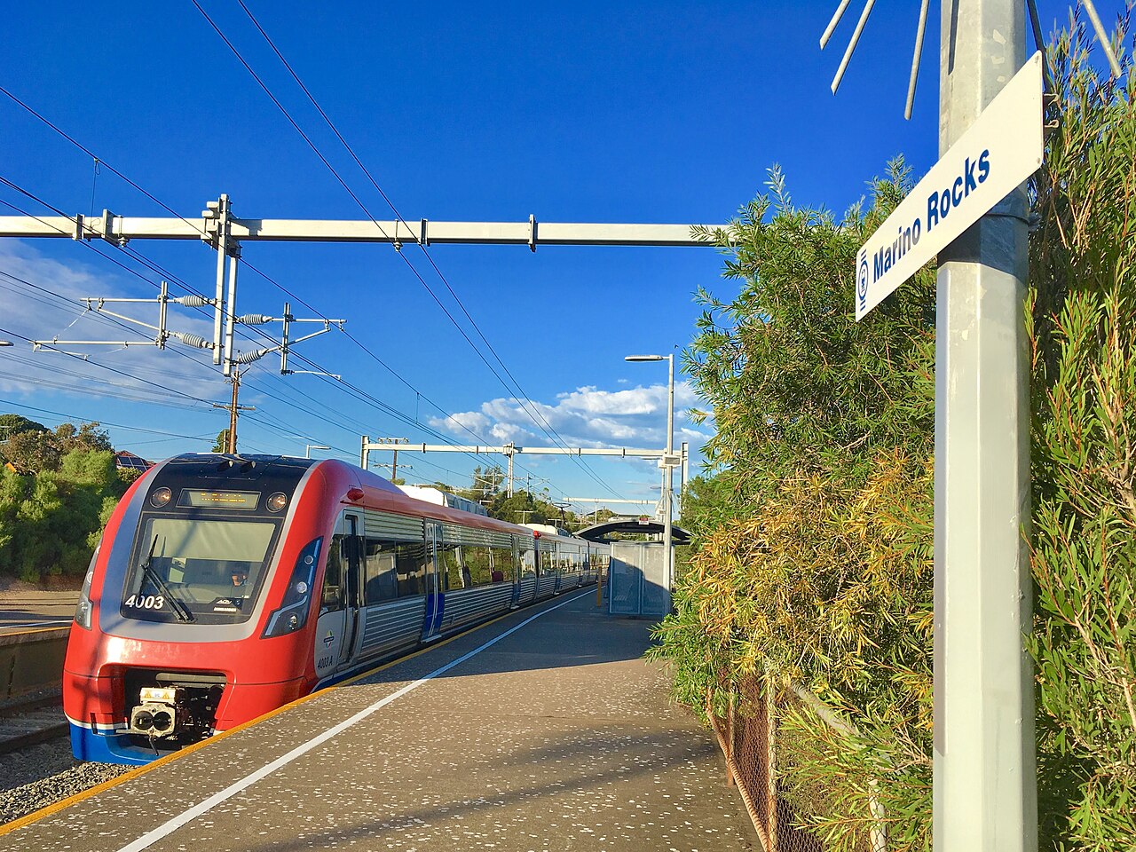 A red, white and blue Adelaide Metro electric train at the platform at Marino Rocks station under a clear blue sky, with the station sign in the foreground