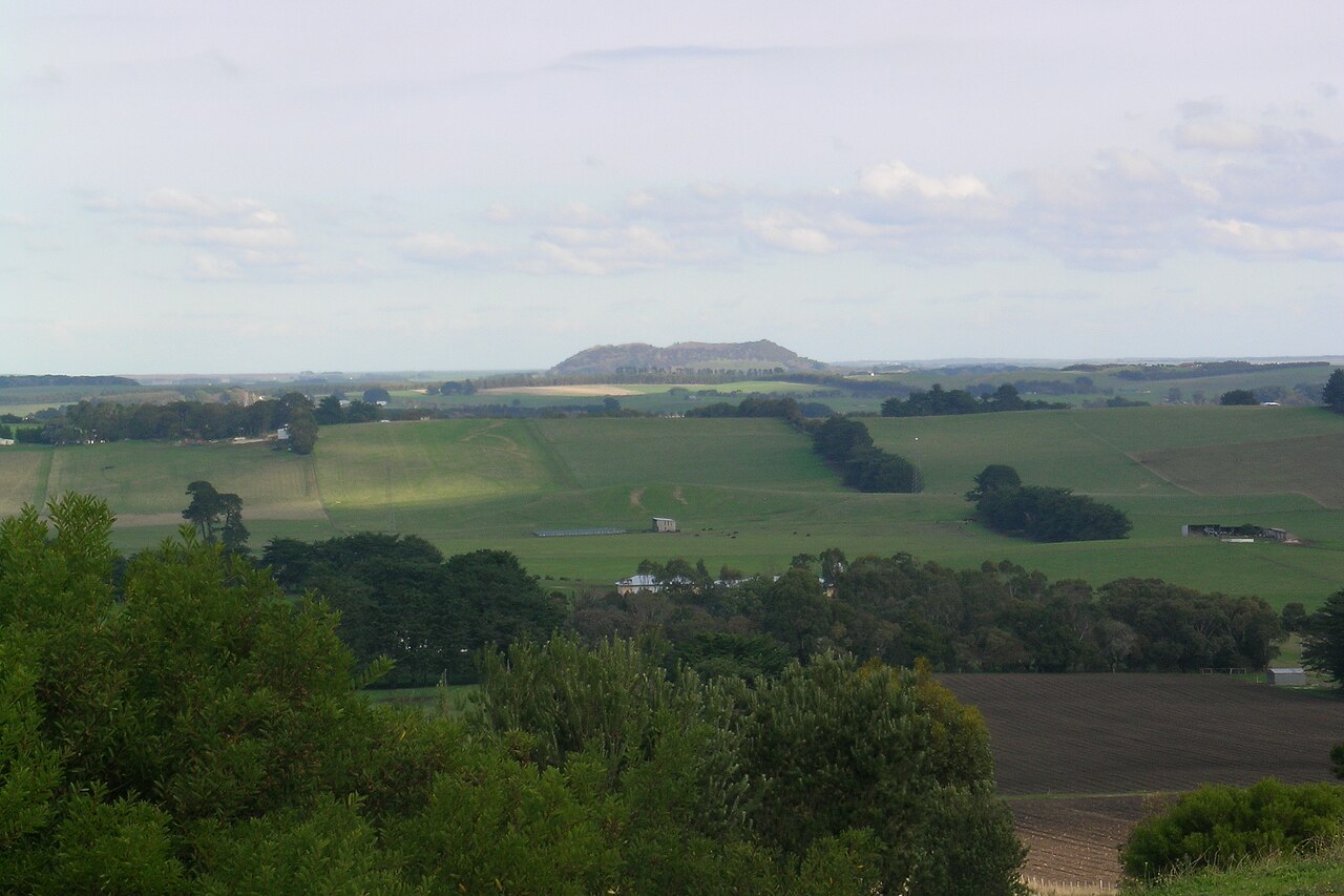 The flat-topped cone of Mount Schank rising above a patchwork of green paddocks and tree lines, seen from a distance under a cloudy sky