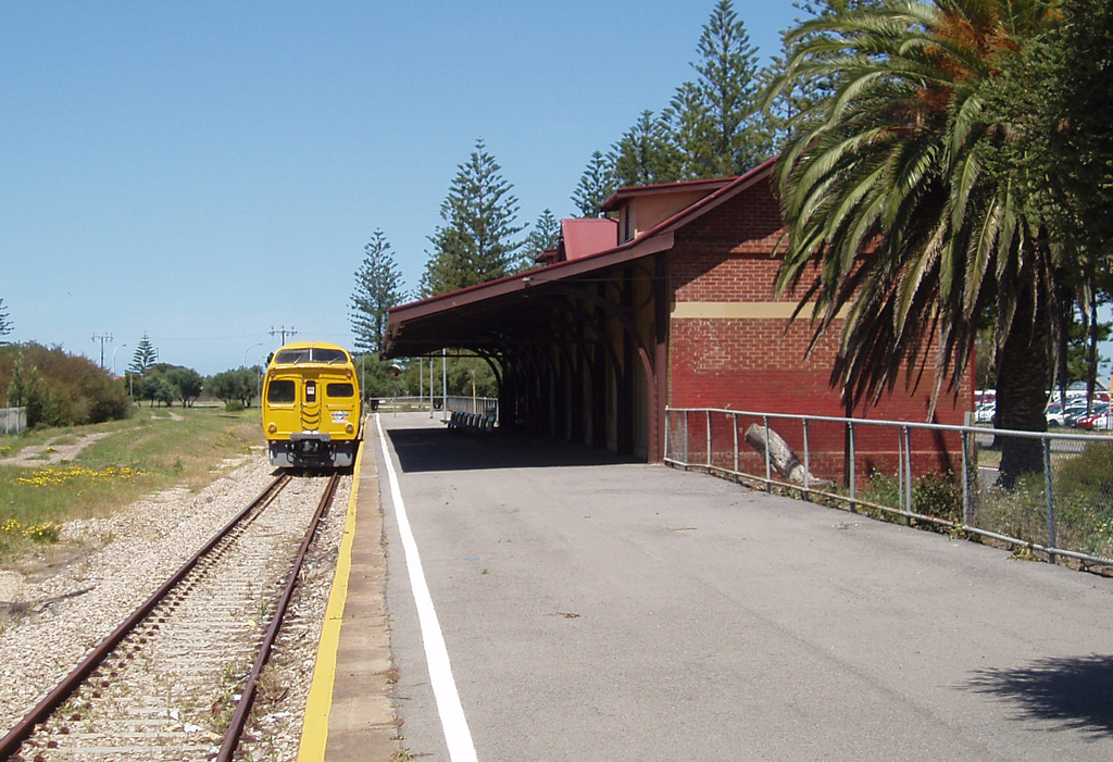 Outer Harbor railway station's red-brick building and long covered platform, with a yellow diesel railcar waiting at the end of the line under a clear blue sky