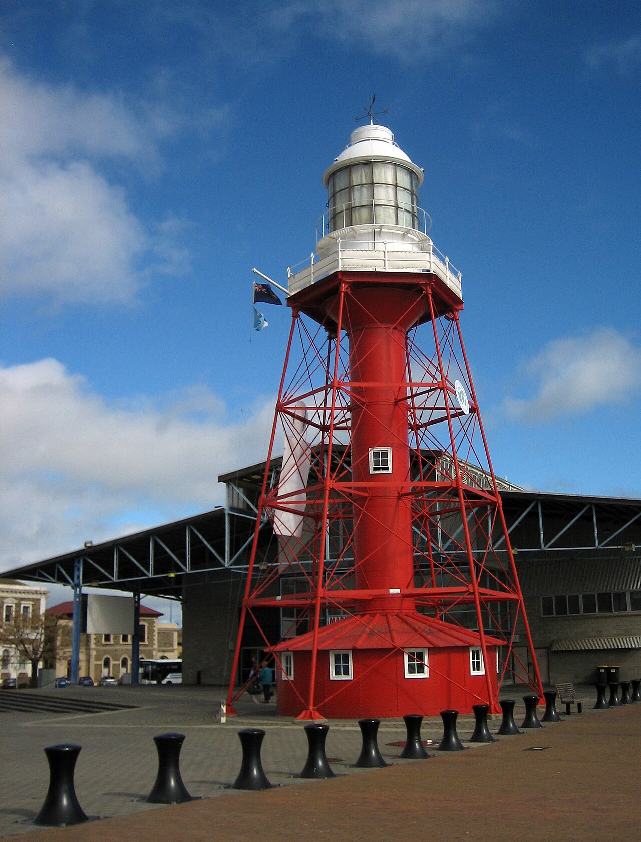 Port Adelaide's red steel lattice lighthouse with a white lantern room, standing on the paved wharf in front of a large open-sided shed, with black bollards in the foreground