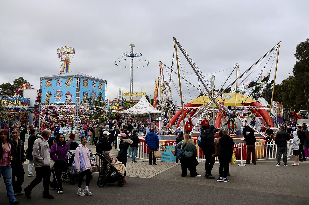 Crowds walking through the Royal Adelaide Show carnival by day, past a bungee trampoline ride, game stalls and a tall swing ride