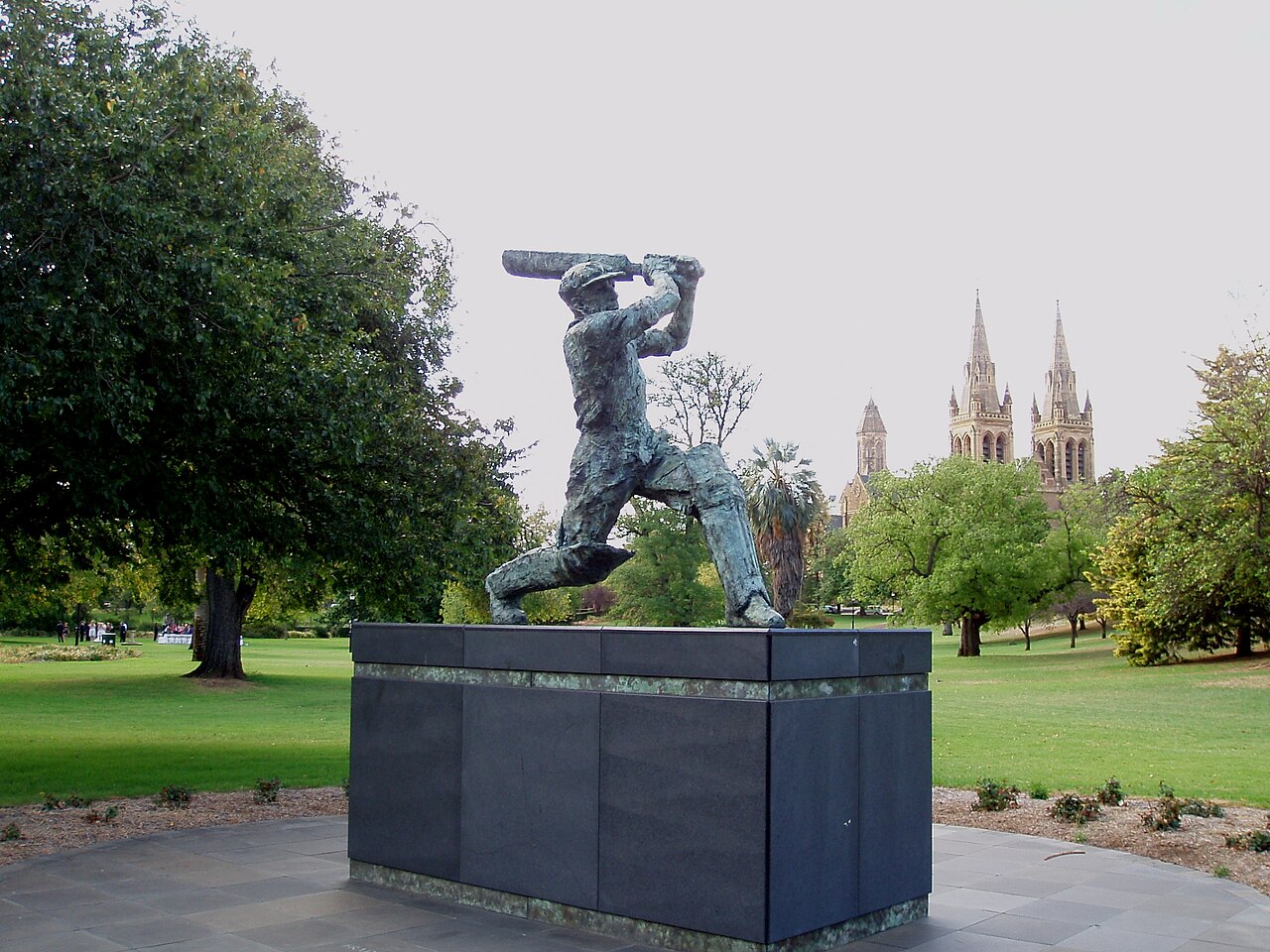 A bronze statue of Sir Donald Bradman mid-stroke on a dark plinth at Adelaide Oval, with trees and the spires of St Peter's Cathedral behind