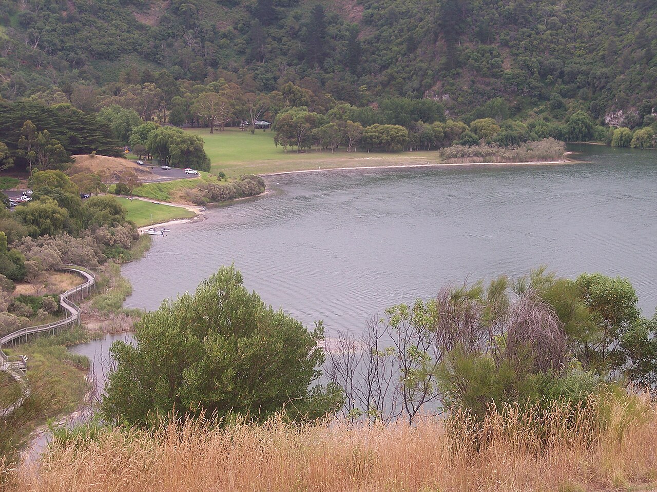 Valley Lake in Mount Gambier seen from above, with grassy picnic areas and trees along the shore, a boardwalk at the water's edge and forested crater slopes behind
