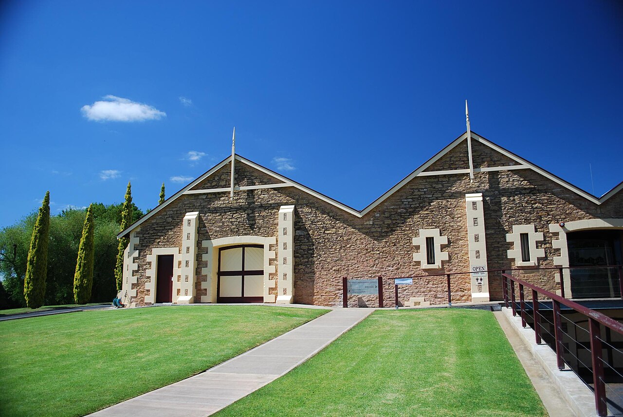 The stone Wynns Coonawarra Estate winery building with three pointed gables, a timber door and a path across a green lawn under a deep blue sky